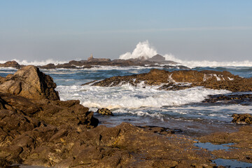 Beautiful landscape with waves beating against brown stones with an old lighthouse and high splashes of the Atlantic Ocean with blue water, white foam waves and a clear clear sky, Portugal