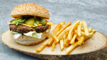 Close-up of juicy cheeseburger with chili pepper, french fries, tomato, lettuce and cucumber pickle on a natural wooden tray with vintage brick wall on background