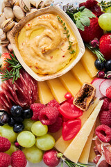 Top view of a cheese board with hummus, fruit and vegetables on a white background