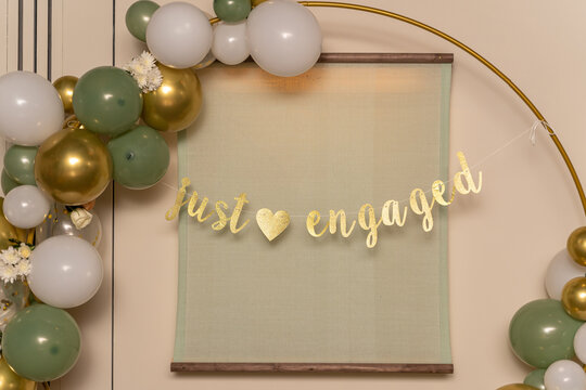 A "just engaged" banner hangs in the middle of an arch adorned with inflated white, green, and gold balloons.