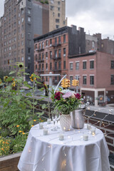 A bouquet of flowers sits on top of a table draped with white tablecloth, candles and fairy lights in celebration of a surprise proposal. Buildings in West Villages are in the background.
