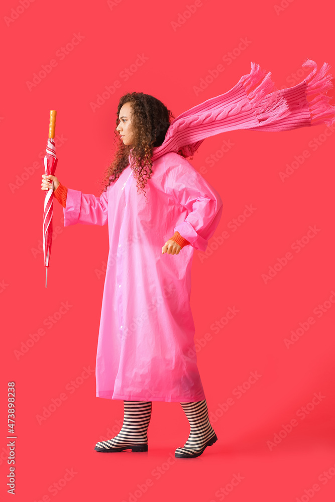 Wall mural Young African-American woman wearing pink raincoat, scarf and gumboots with umbrella on red background