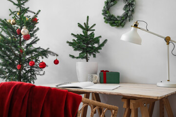 Comfortable workplace with lamp, opened notebook, Christmas tree and gift box near light wall