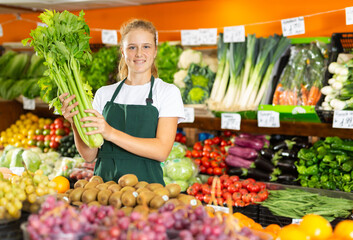 Obraz premium Portrait of positive teenage girl in uniform working in grocery shop as job experience, selling celery