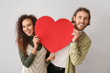 Happy young couple with red paper heart on light background. Valentine's Day celebration