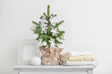 Beautiful Christmas tree in pot decorated with snowflakes, balls, blank frame and warm clothes on table near white wall
