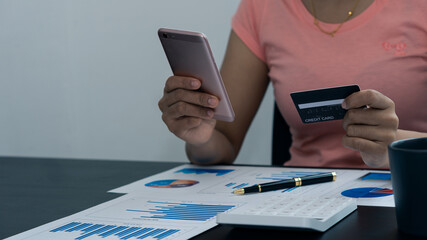 Young woman holding phone and credit card with money chart and pen on the table on an online business concept.