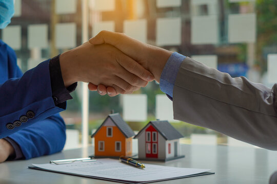 A young man shakes hands with real estate agents in offices and houses in the background and offers a price list and terms of purchase or rent on his workspace table.