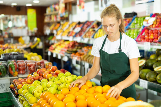 Hardworking fifteen-year-old girl who works part-time in a store as a trainee saleswoman puts oranges on the counter