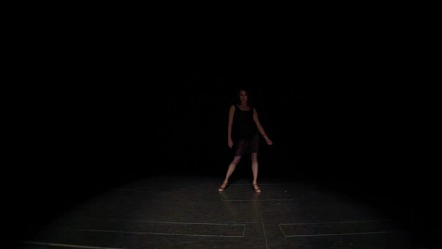Woman ballroom dancer comes out of darkness onto marked stage with slow,flexible steps in Latin American tango dance style,turns around,stumbles,stroking,flexing body passionately.Black dress,leotard