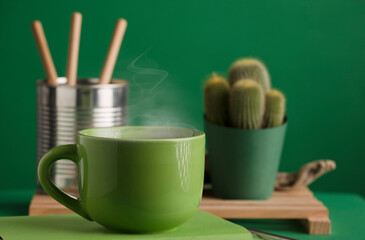 cup of green tea or coffee, hot with steam, cactus and recycled metal can, green background.