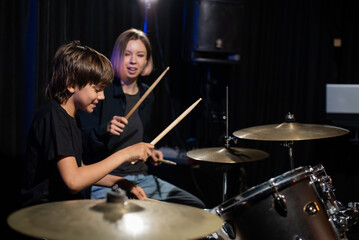 Young caucasian woman teaches a boy to play the drums in the studio on a black background. Music...