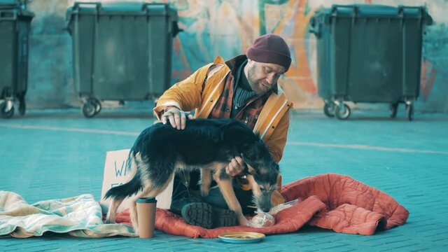 A tramp is petting his dog while sitting on the ground