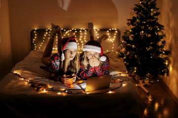 Two teenage girls in plaid shirts lying on the bed and using a laptop, a lighted Christmas tree in...