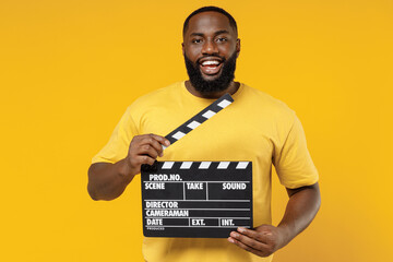 Young cheerful fun happy black man 20s wearing bright casual t-shirt holding classic black film making clapperboard isolated on plain yellow color background studio portrait. People lifestyle concept.