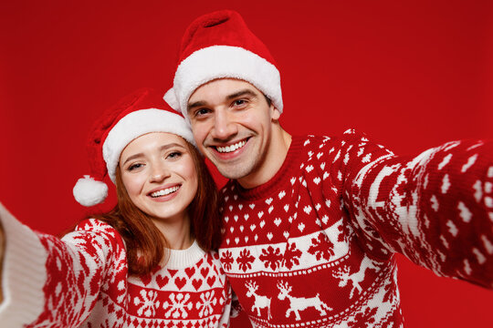 Close up young couple friends two man woman 20s in sweater hat doing selfie shot pov on mobile phone isolated on plain red background. Happy New Year 2022 celebration merry ho x-mas holiday concept.