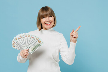 Elderly woman 50s in white knitted sweater holding fan of cash money in dollar banknotes point index finger aside on copy space isolated on plain blue color background studio People lifestyle concept