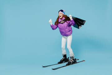 Full body skier woman in warm purple padded windbreaker jacket ski goggles mask spend weekend in mountains holding package bags with purchases after shopping isolated on plain blue background studio
