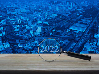 Magnifying glass with 2022 text and financial graph on wooden table over modern office city tower, street, expressway, and skyscraper, Business happy new year 2022 research concept