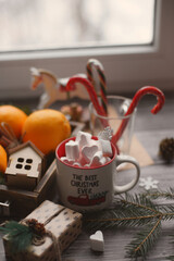 A mug with marshmallows, caramel canes, toys, oranges in a box stand on a light background near the window.