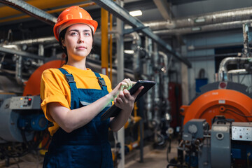 Concept of industrial production and equality. Portrait of a caucasian woman-worker in uniform and helmet with a tablet in her hand. In the background boiler room