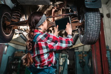 Obraz na płótnie Canvas A young female mechanic, in uniform, holds a tablet in her hands and inspects the car. Car on the lift. Rear and bottom view