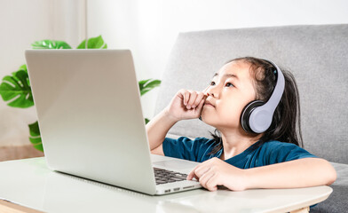 A cute child girl thinks has an idea while online studies on a laptop at home due to the Covid-19 coronavirus pandemic. Online education learning gaming technology and homeschooling concept idea