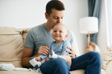 Affectionate father kisses his baby son who is sitting on his lap at home.