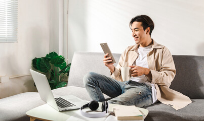 Happy smiling Asian student sits and relaxes on a couch using modern tablet browsing unlimited wireless internet, positive young man freelancer working uses wifi at a home apartment, technology idea