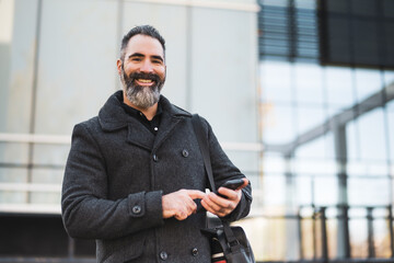 Portrait of happy businessman in black clothing standing in front of company building. Black bearded man is wearing winter clothing and enjoying using smartphone.