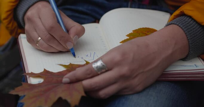 Close up hands of woman sitting on bench in autumn park. She writes formulas using pen and uses yellow oak leaf as bookmark and closes notebook. ���������� - september