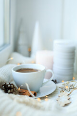 A cup of coffee over Christmas bokeh garlands in the house on the table with a sweater in the background and decorations. Winter mood, festive decoration, magical Christmas.