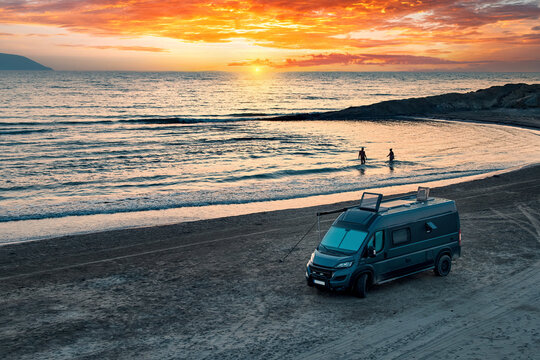Aerial photo of campervan on abandoned beach against beutiful sunset. People bathing in the sparkling sea. Outdoor nomad lifestyle, van life holiday. Independent road trip concept.