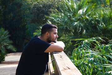 A gay man of Latin origin, dark and bearded, stands pensively looking nowhere in the park. He is sad, dazed and depressed. Concept of expressions of sadness and depression.