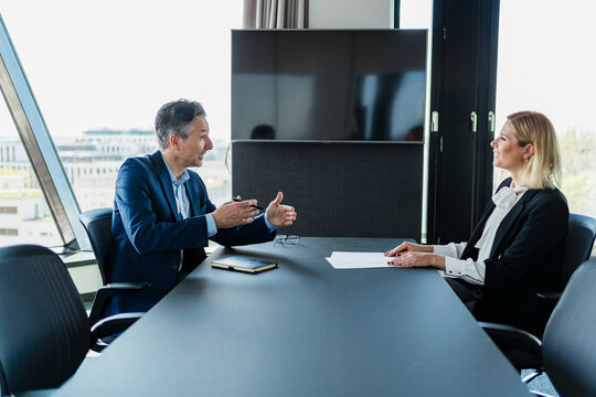 Mature businessman gesturing while discussing with female coworker sitting in board room