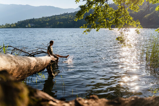 Man dangling legs and splashing water while sitting on fallen tree at lakeshore