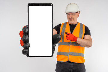 Builder with big phone. Place to advertise construction and repair work on smartphone screen. Master holds out phone and points at its screen. A construction worker in a helmet and a reflective vest.
