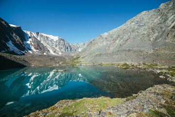 Fototapeta na wymiar Snowy mountain reflected in clear water of glacial lake. Beautiful sunny landscape with glacier reflection in water surface of mountain lake under clear sky. Snow on rock reflected in mountain lake.