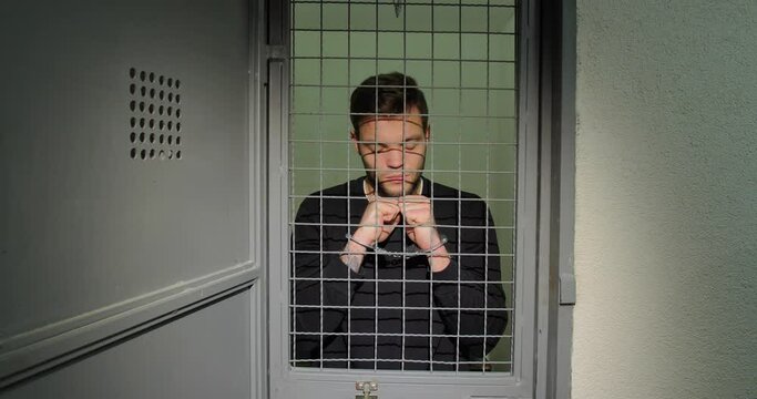 A man in handcuffs in a prison cell stands upset holding to a lattice. Offender.