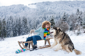 Boy with dog enjoy a sleigh ride. Child sledding, riding a sledge. Children play in snow in winter. Outdoor kids fun for Christmas vacation.