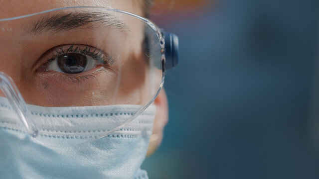 Scientist showing one eye on camera in laboratory, wearing safety glasses and face mask. Woman biologist looking at camera with face of face, working in research lab. Close up