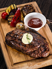 grilled beef angus steak with melted butter on a wooden cutting board served with grilled vegetables, close up photo on a dark background