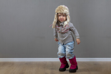 Cute baby girl 2 years old caucasian in warm winter clothes and a hat with earflaps with fur on a gray background