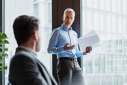 Mature businessman with documents gesturing while talking to colleague in office