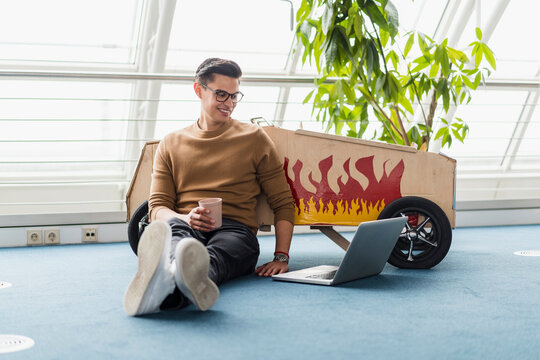 Male professional smiling while sitting by laptop on toy car in creative office