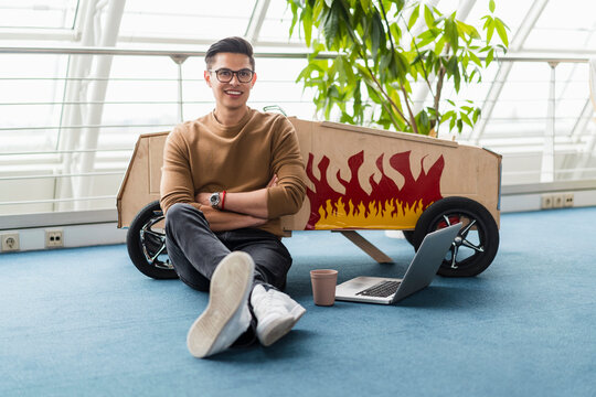 Confident creative businessman with arms crossed sitting by laptop and toy car in office