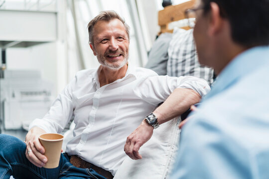 Smiling male professional holding disposable cup while sitting with colleague at office