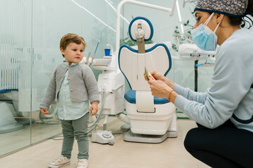 Toddler boy looking at female pediatric dentist with protective face mask in clinic