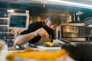 Male chef in protective face mask garnishing food in kitchen at restaurant