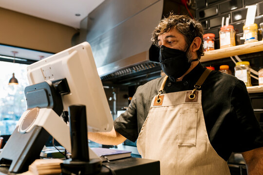 Mature male cashier working on computer at counter in restaurant during pandemic